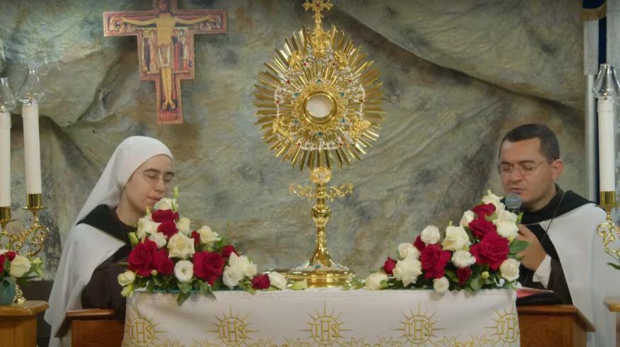 Fotografia de dois sacerdotes, uma mulher e um homem, rezando diante de uma imagem dourada