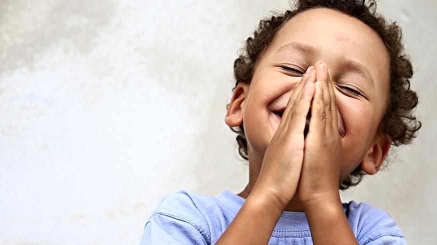 Fotografia de um menininho de pele negra-clara com as mãos fechadas em oração, sorrindo de olhos fechados